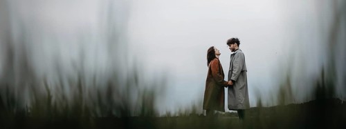A couple standing apart in a field on an overcast day, holding hands, conveying a sense of