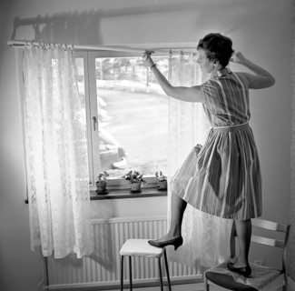 1960s home decorating. A woman at home putting up the curtains in the room window. All by herself