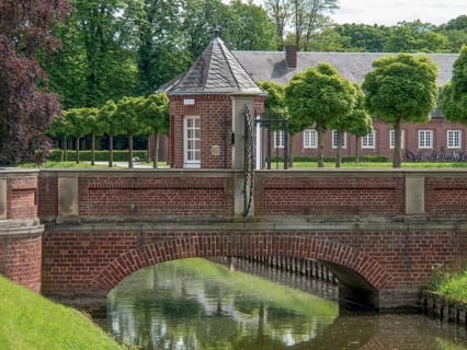 A brick bridge over a body of water, surrounded by manicured hedges and a castle complex,