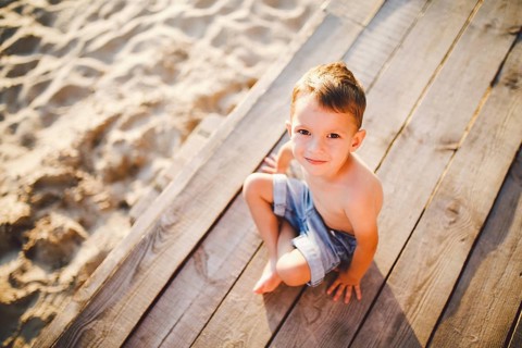 The theme is a child and summer beach holiday. A small Caucasian boy sits sideways on a wooden pier