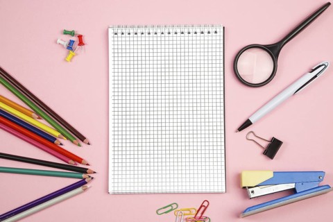Colored pencils, stationery, paper notebook with pen and magnifier on pink isolated background. top