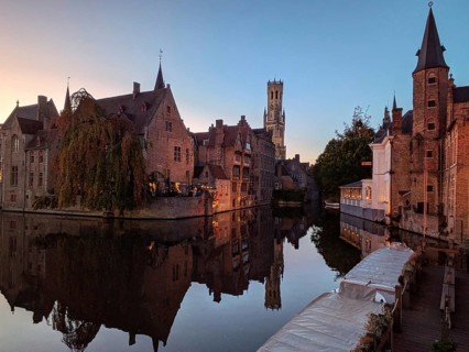 Charming canal at sunset with medieval buildings and a distant tower reflected in the calm water.