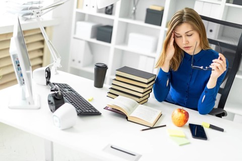 A young girl is sitting at a computer desk, holding glasses in her hand and rubbing her eyes.
