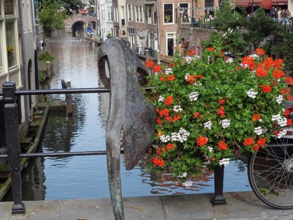 Sculpture and bicycle on a railing along a canal with blooming flowers and reflecting buildings,