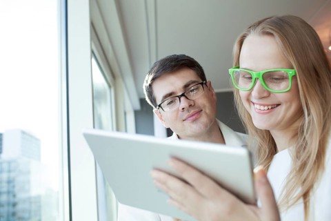 Man and woman in glasses are standing by window and watching something in tablet PC