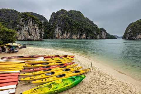 Several colourful kayaks lying on the beach in front of green rocks and calm water under a cloudy