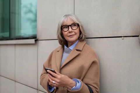 Eine reife Frau in einem hellbraunen Mantel steht an einem modernen Gebäude und überprüft ihr
