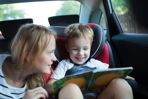 Little boy in a car sitting in child safety seat looking at the pictures in the book, mother