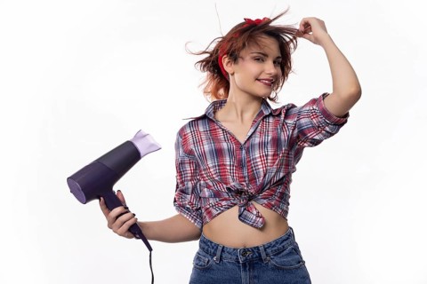 A playful young woman joyfully uses a hair dryer, delighting in her hairstyle with vibrant energy