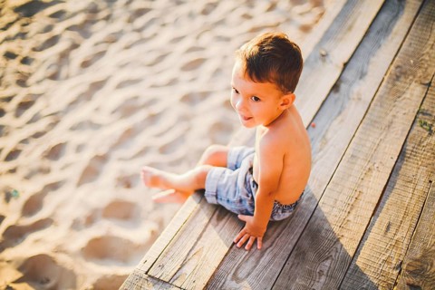 Little Caucasian boy child sitting on wooden pier sandy beach, summer time, sea holiday near water.