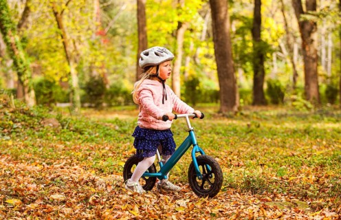 Side view preschool girl in protective helmet riding blue balance bike at the autumn park. Lovely