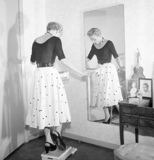 1950s fashion. A young woman is standing on one leg on the scale trying to be as light as possible.