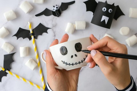 Female hands paint on white cup scary jacks face DIY for kids Halloween home activities Holiday art