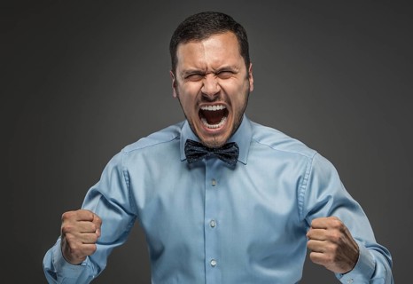 Portrait of angry upset young man in blue shirt and butterfly tie with fists up yelling isolated on