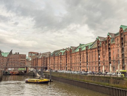 Yellow boat on a river next to historic brick-red buildings and a metal bridge under a cloudy sky,