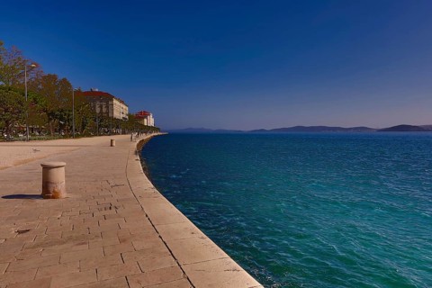 Long promenade along the blue water, with historic buildings and a clear sky in the background,