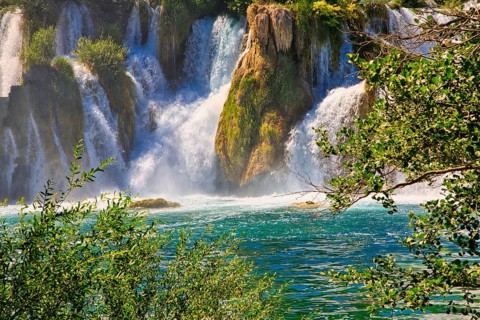 An impressive waterfall flows into a clear, green river, surrounded by lush vegetation and bright