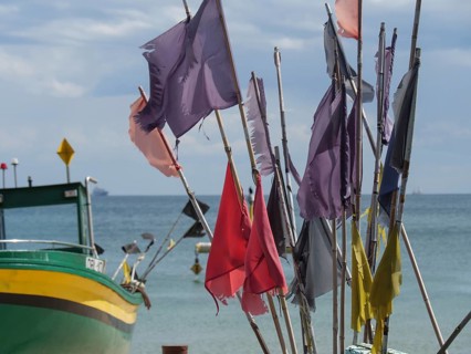 Colourful flags waving in the wind, next to a green boat in front of a wide sea and blue sky,