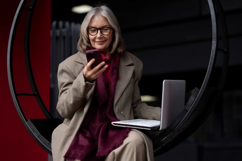 A senior businesswoman engages in work on her laptop outside, occasionally checking her smartphone.