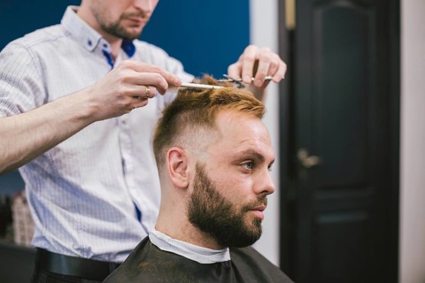 Barber Making Haircut Bearded Man In Barbershop. Professional stylist cutting client hair in salon.