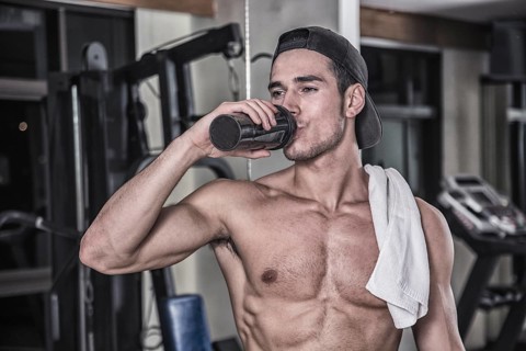 Attractive athletic shirtless young man drinking protein shake from blender in gym while looking at