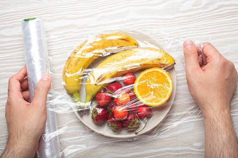 Male hands wrapping assorted different fruit and berries on white plate in plastic clear film top
