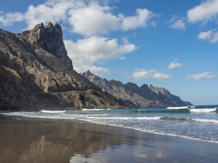 A peaceful beach with rocky cliffs and gently breaking waves under a blue sky and white clouds,