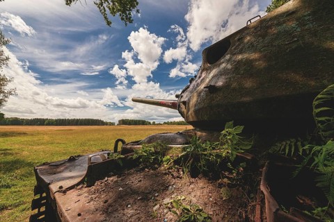 An abandoned tank stands in a meadow under a cloudy sky, surrounded by open and green nature in