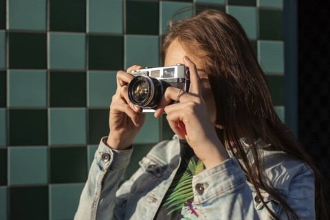 Cool girl model with retro film camera wearing a denim jacket, dark hair outdoors over city wall in