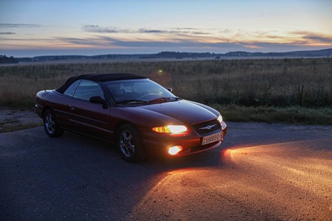 Convertible car parked on a road at sunset, casting an orange glow with fields around