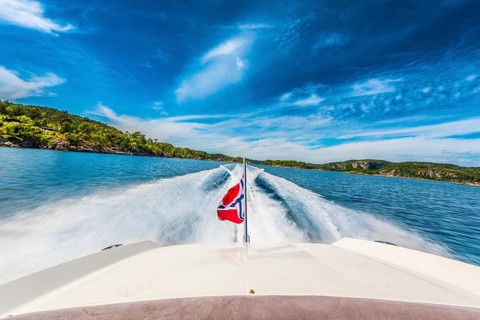 Speed boat swiftly crossing a sunny fjord in southern Norway sporting the Norwegian flag.