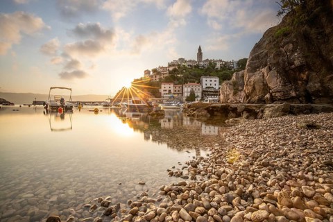 Harbour and beach with an old village on a hill. Boats are reflected in the water, the sun rises on