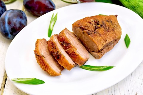 Roasted duck breast with green onions in a white oval plate, towel and plums on a background of