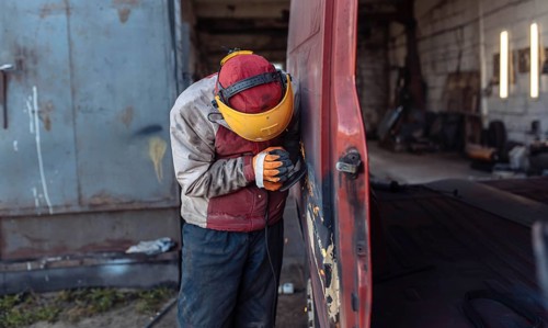 A worker sands a rusty van in an outdoor workshop setting