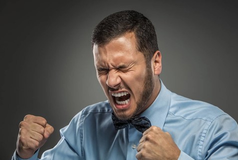 Portrait of angry upset young man in blue shirt and butterfly tie with fists up yelling isolated on