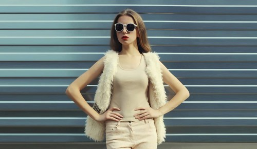 Portrait of modern stylish young woman in white fur vest, glasses, fashionable female model posing