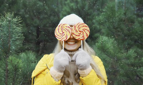 Happy funny smiling young woman having fun with lollipop enjoys snow, in winter park against