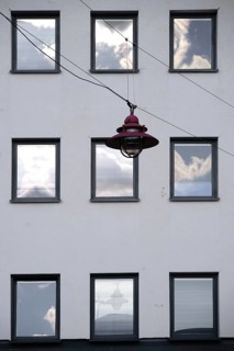 A hanging lamp in front of the façade of a residential building with windows reflecting the clouds