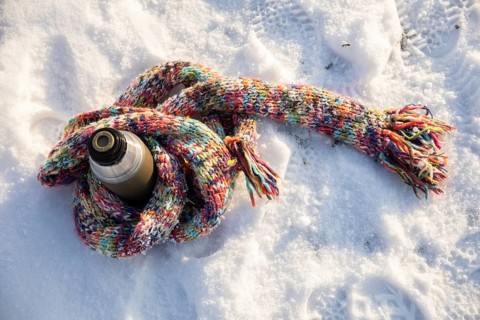 A long, colorful wool scarf lies on the snow. The scarf is wrapped around a thermos. The thermos is