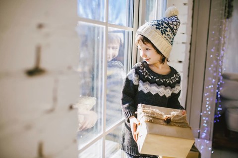 Child sitting at the window on a sunny Christmas day, making presents wrapped in boxes in paper.
