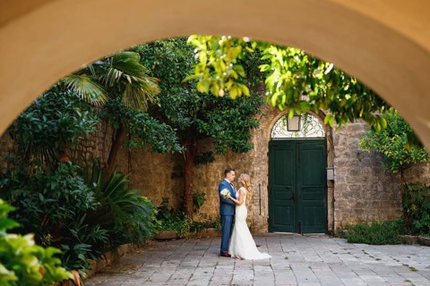 The bride and groom stand hugging in the coutryard in the old town of Perast, view through the arch