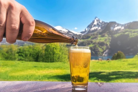 Hand of a man pouring beer from a bottle in a glass with a summer scenery in the background with