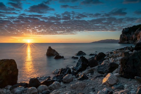 A colorful sunrise at Cala Gonone with black and white rocks and boulders and a sunburst