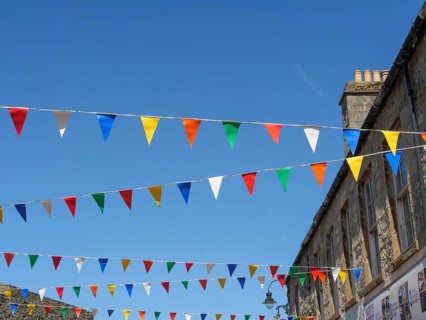 Colourful flags hang over a street corner between historic buildings under a cloudless blue sky,