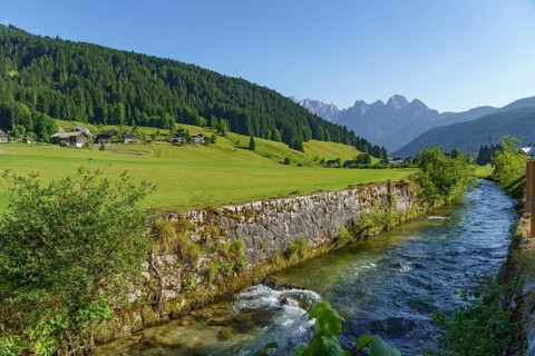 A clear river flows between wide green meadows and a mountain landscape under a bright blue sky,