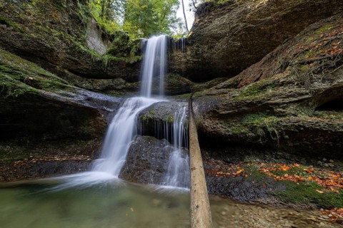A waterfall flows over moss-covered rocks in a secluded forest, covered with autumn leaves,