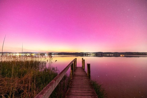A wooden footbridge leads over a calm lake under a sky with aurora borealis, Reichenau, Lake