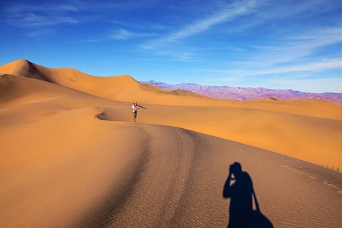 Orange sand dunes in Death Valley, California. A middle-aged woman in striped T-shirt carries a