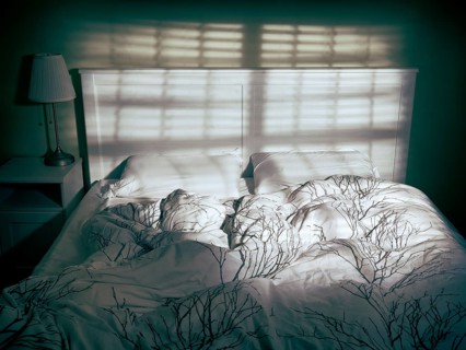 Unmade empty bed in dramatic dim light coming from a window through blinds