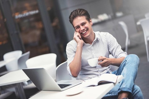 Handsome caucasian young man with phone working on laptop and smiling while enjoying coffee in cafe